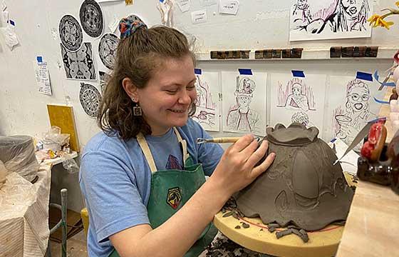 Emily seated before upside down piece of pottery, carving, and smiling