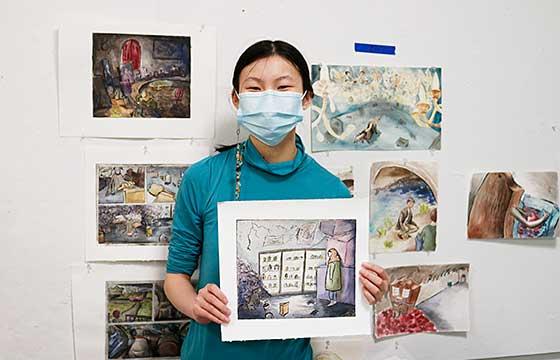 Ting standing in studio, mask on, holding small painting with a backdrop of other paintings hung on walls