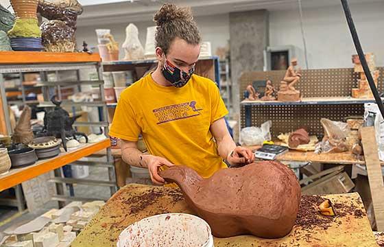 Vincent in studio wearing yellow AU Simming shirt, carving large terra cotta sculpture resting on a piece of foam